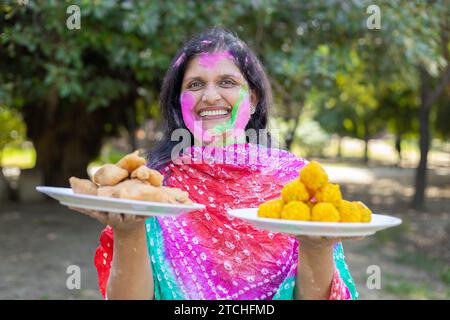 Mujer india feliz que lleva kurta blanca sosteniendo platos de gujiyas y laddus. Celebrando el festival holi en el parque o el jardín. Foto de stock