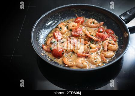 Camarones en salsa de crema de tomate salteados con hierbas, cebollas y ajo en una sartén negra en la estufa, cocinando una comida de marisco mediterráneo, sele Foto de stock