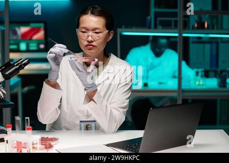 Joven investigador asiático examinando atentamente partículas de carne cruda mientras trabajaba en laboratorio Foto de stock