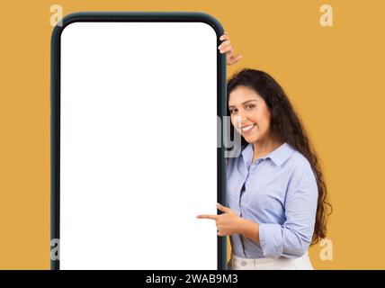 Mujer joven alegre en una camisa azul se asoma desde detrás de un gran smartphone Foto de stock