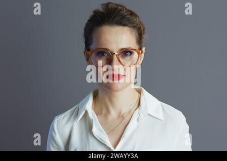 mujer elegante de mediana edad del dueño de la pequeña empresa en blusa blanca con gafas aisladas en gris. Foto de stock