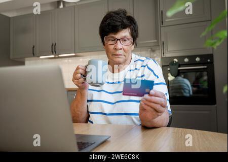 Mujer mayor que hace el pago de la tarjeta de crédito a través del ordenador portátil en la cocina Foto de stock