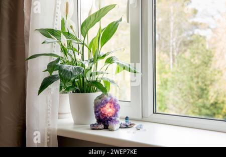 Lámpara de geoda amatista iluminada, atmósfera de hogar calmante espiritual. Planta de limpieza de aire flor Spathiphyllum, spath o lirio de la paz que crece en la ventana. Foto de stock