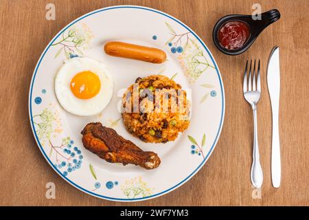 Arroz frito americano con palo de tambor frito, huevo frito y salchicha en plato de cerámica blanca con ketchup en taza negra, tenedor de metal y cuchillo en mesa de madera Foto de stock