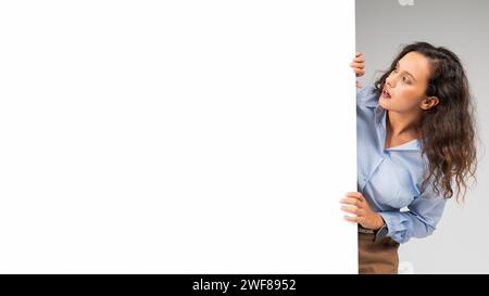 Curiosa y atenta, una mujer joven con el pelo rizado se asoma detrás de una pared blanca en blanco Foto de stock