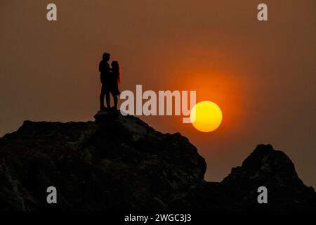 Baluchistán, Pakistán - 16 de enero de 2024: Siluetas pareja de pie en la roca en la playa con la puesta de sol en el fondo. Foto de stock