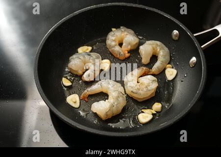 Los camarones pelados crudos y el ajo se saltean en una sartén en la estufa negra, preparación para un delicioso aperitivo, espacio de copia Foto de stock