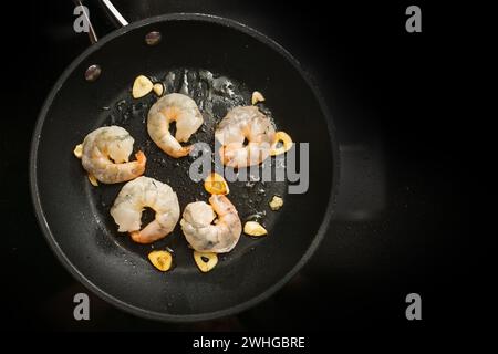 Los camarones frescos se saltean con ajo en aceite de oliva en una sartén en la estufa negra, preparación para un delicioso aperitivo, c Foto de stock