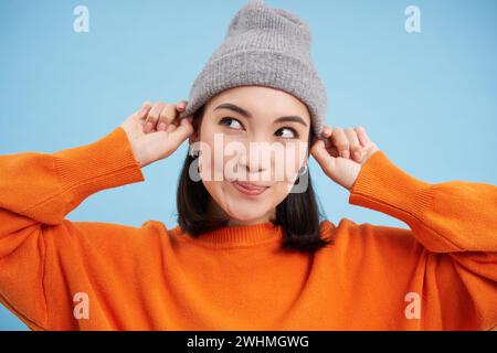 Close up retrato de la muchacha coreana feliz, elegante pone el sombrero caliente, sonríe y se ve alegre, se coloca contra el fondo azul del estudio Foto de stock
