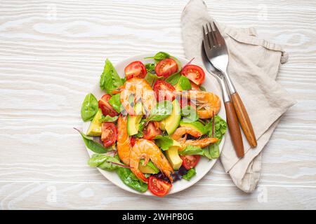 Ensalada saludable con camarones a la parrilla, aguacate, tomates cherry y hojas verdes en plato blanco con cubiertos en madera blanca rústica Foto de stock