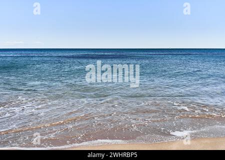 Un paisaje marino tranquilo y claro con aguas azules que chapotean suavemente sobre una orilla arenosa bajo un vasto cielo azul. Foto de stock