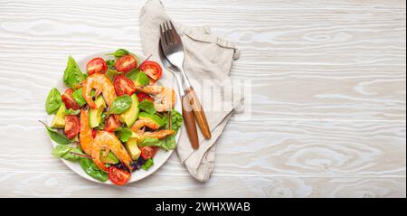 Ensalada saludable con camarones a la parrilla, aguacate, tomates cherry y hojas verdes en plato blanco con cubiertos en madera blanca rústica Foto de stock