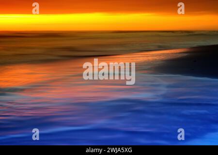 Una pacífica puesta de sol arroja vibrantes tonos anaranjados y azules sobre el horizonte del océano y suaves olas. Foto de stock