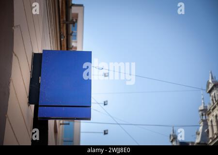 Un letrero cuadrado azul cobalto vacío y vibrante, exhibido prominentemente y colgando de un soporte de metal contra una fachada del edificio, captura la esencia Foto de stock