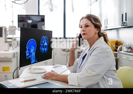 Médico femenino que trabaja en la computadora en el consultorio del médico, mirando la resonancia magnética, llamando por teléfono los resultados de la prueba al paciente. Doctor consultando escaneo con la othe Foto de stock