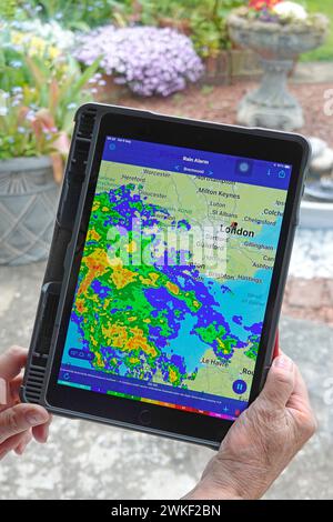 Las manos de la mujer sostienen la aplicación de alarma de lluvia de la tableta Apple ipad pro y la capa de datos del radar del color en tiempo real del mapa local de la lluvia tiempo húmedo en la ventana de casa Essex UK Foto de stock
