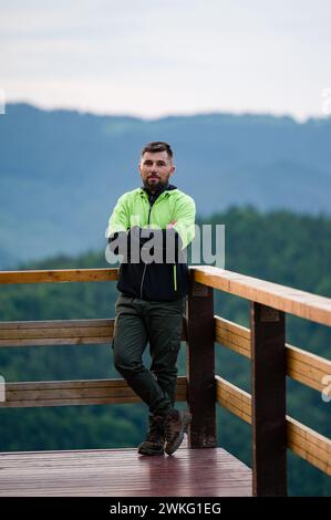 un hombre con ropa deportiva se encuentra apoyado en una barandilla de madera en una terraza de madera, turista en las montañas de vacaciones. Foto de stock