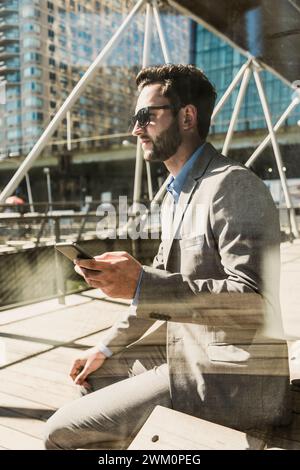 Hombre de negocios que sostiene el teléfono inteligente sentado en el banco en la ciudad en el día soleado Foto de stock