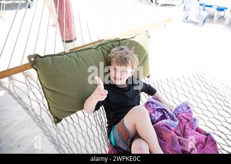 Niño joven sonriente dando pulgares para arriba mientras está sentado en la hamaca en la playa Foto de stock