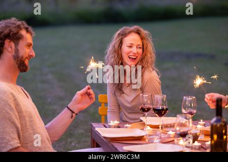 La imagen captura una escena conmovedora de un hombre y una mujer disfrutando de un momento lleno de risas con chispas en una fiesta de vino rústica en el jardín durante el crepúsculo. Noche alegre con Sparklers en una reunión de vino en el jardín. Foto de alta calidad Foto de stock