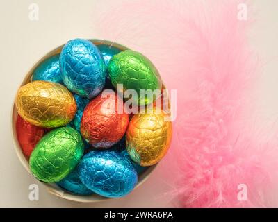 Tazón de madera lleno de papel cubierto de chocolate huevos de Pascua con plumas rosadas Foto de stock
