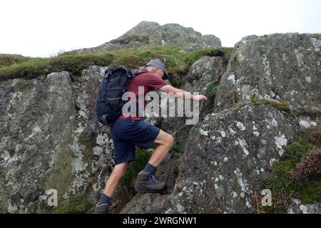Hombre (excursionista) en pantalones cortos con mochila que sube a las rocas en el Wainwright 'Arnison Crag' en Patterdale, Lake District National Park, Cumbria, Inglaterra, Reino Unido Foto de stock