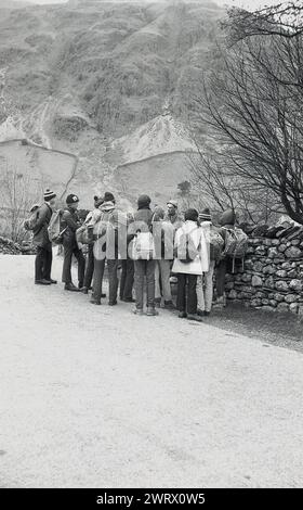 1970s, un grupo de jóvenes en un curso de orientación exterior, de pie junto a una pared de piedra escuchando a su instructor, Ambleside, Lake District, Cumbria, Inglaterra, REINO UNIDO. Foto de stock