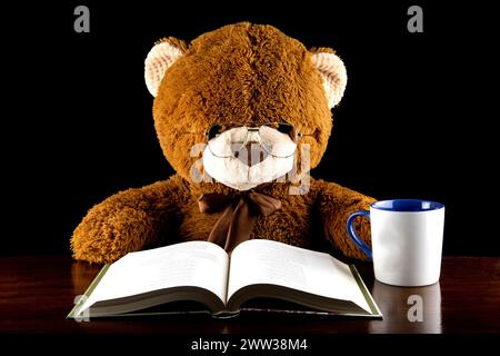 Oso de peluche marrón grande sentado en una mesa de lectura con una taza de café o té Foto de stock