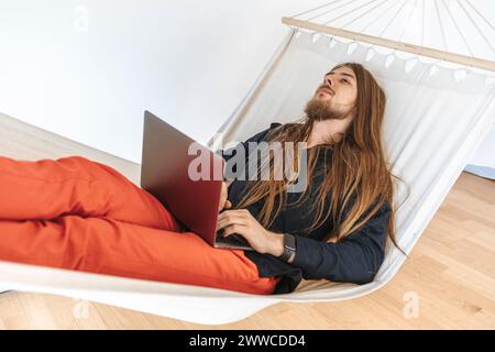 Programador joven que se relaja en la hamaca con el ordenador portátil en el inicio en la oficina Foto de stock