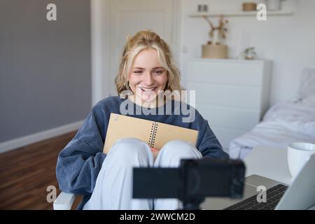Retrato de mujer rubia sonriente, graba video en cámara digital cómo escribe en cuaderno, habla con seguidores, haciendo contenido de blog de estilo de vida en su r Foto de stock