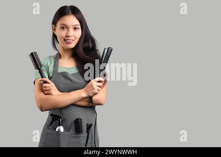 Hermosa peluquería femenina asiática con cepillo y alisado de hierro sobre fondo gris Foto de stock