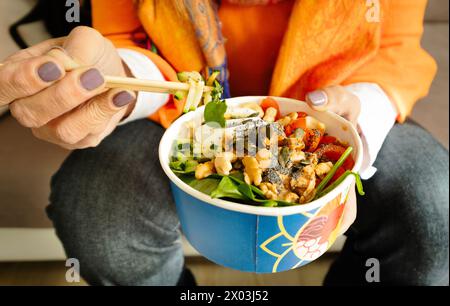 Primer plano de las manos de una mujer sosteniendo un tazón colorido de ensalada asiática (Poke) con palillos Foto de stock