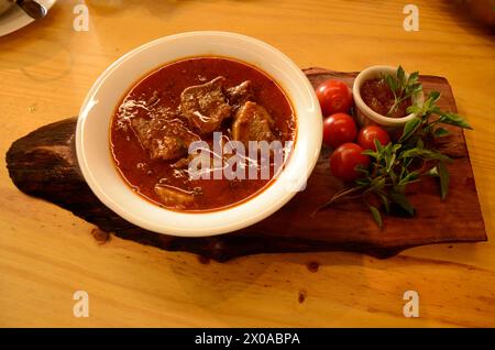 En una tabla de madera, un tazón pequeño blanco con lengua de ternera en salsa, tomates cherry, albahaca y un recipiente con salsa de pimienta y ramitas de menta. Foto de stock
