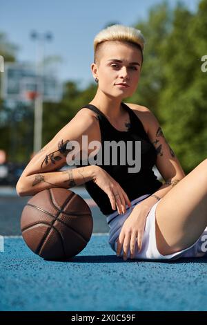 Una mujer joven con tatuajes se sienta en el suelo, sosteniendo un baloncesto, perdido en el pensamiento. Foto de stock