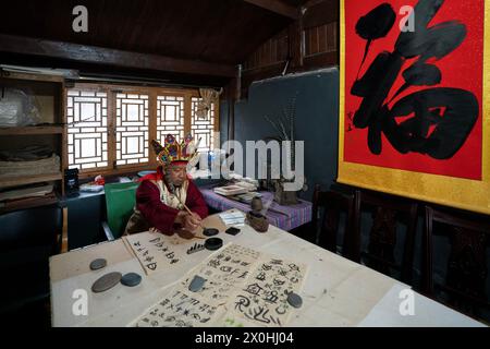 (240412) -- LIJIANG, 12 de abril de 2024 (Xinhua) -- He Jielin escribe en la pictografía de Dongba en casa en la aldea de Yuhu, ciudad de Lijiang, provincia de Yunnan, al suroeste de China, 25 de marzo de 2024. Viviendo en la aldea Yuhu al pie de la montaña de nieve Yulong, Jielin ha sido testigo de los cambios en esta antigua aldea Naxi. Desde los años 1980 hasta los años 1990, Jielin y su esposa solo podían vivir de la tierra. Al igual que otros aldeanos, la pareja se ganaba la vida cortando madera, recogiendo materiales medicinales y cavando arena, lo que resultó en grandes daños al bosque y al medio ambiente. Sin embargo, la gente en el pueblo permanece Foto de stock