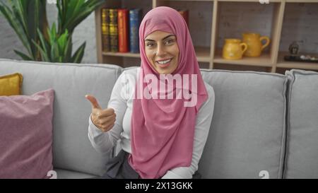 Mujer sonriente en hijab dando pulgares para arriba en el interior en el sofá acogedor de la sala de estar Foto de stock