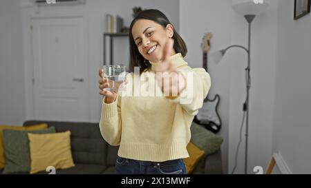 Mujer hispana joven sonriente con un vaso de agua dando pulgares para arriba en un ambiente acogedor de la sala de estar. Foto de stock