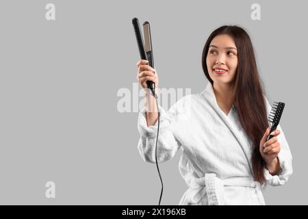 Mujer asiática feliz joven hermosa en albornoz con cepillo y hierro enderezado sobre fondo gris Foto de stock