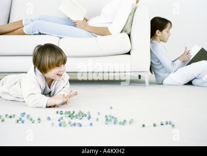 Niño jugando canicas en el suelo, la mujer y la niña leyendo en segundo plano.