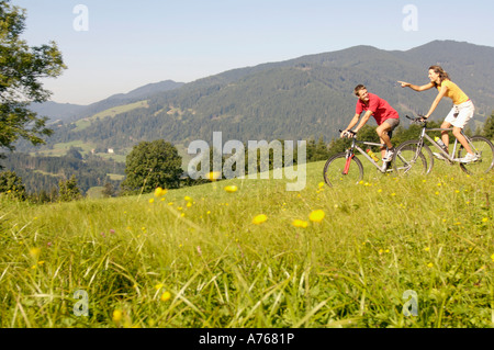 Pareja joven montando en bicicleta de montaña en la pradera, vista lateral, con montañas al fondo Foto de stock
