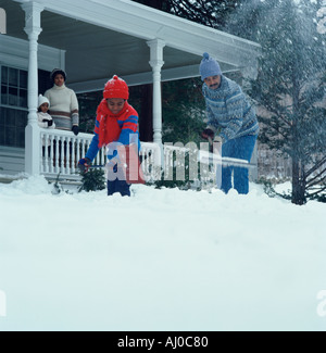 Padre e hijo afroamericano palear la nieve de su pasarela como madre e hija mira desde el porche Foto de stock