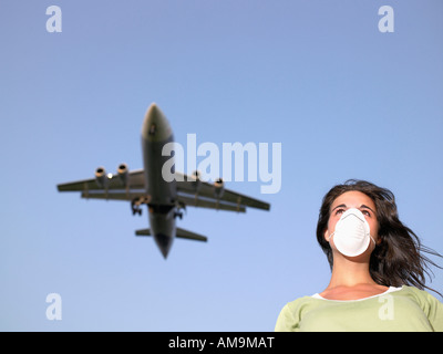 Mujer vistiendo una máscara quirúrgica mientras un avión vuela encima. Foto de stock