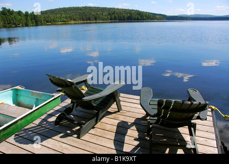 Dos sillas de madera Adirondack en el muelle frente a un lago azul con nubes reflexiones Foto de stock