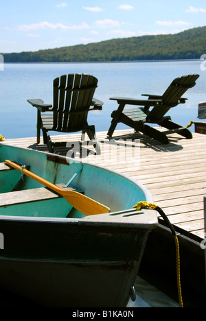 Botes y dos sillas de madera Adirondack en el muelle frente a un lago azul Foto de stock