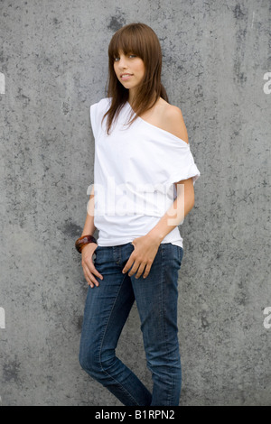 Joven, mujer morena con jeans y un top blanco, de pie delante de una pared gris, relajado, mirando a la cámara Foto de stock