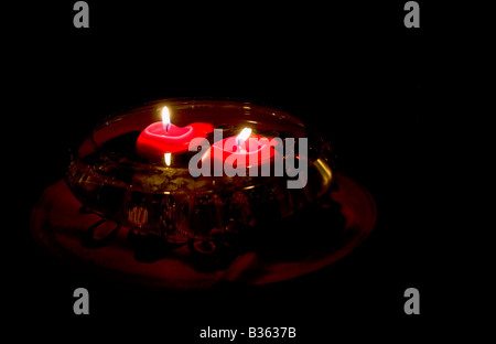 Un recipiente contiene agua y dos velas rojas flotantes con una llama en la oscuridad. Foto de stock