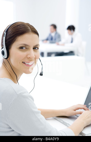 Mujer vistiendo auriculares, trabajando en un escritorio, sonriendo a la cámara sobre el hombro Foto de stock