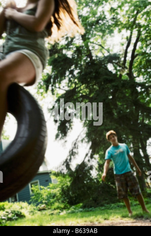 Adolescente balanceándose en un columpio de neumático con un joven en el fondo Foto de stock