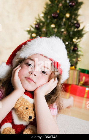 Retrato de niña (4-5) en Santa hat con osito de peluche, árbol de Navidad y se presenta en segundo plano.