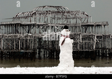 Un muñeco de nieve en hat bufanda y gafas de sol en la playa de Brighton en frente del esqueleto del Old West Pier Foto de stock
